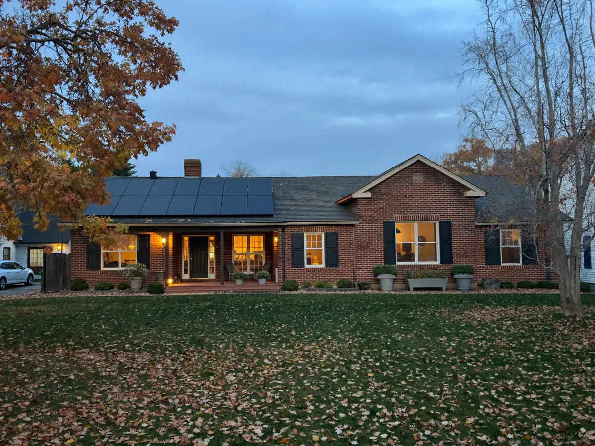 A brick ranch home with solar panels on its roof at dusk, porch light on and autumn leaves on the lawn.