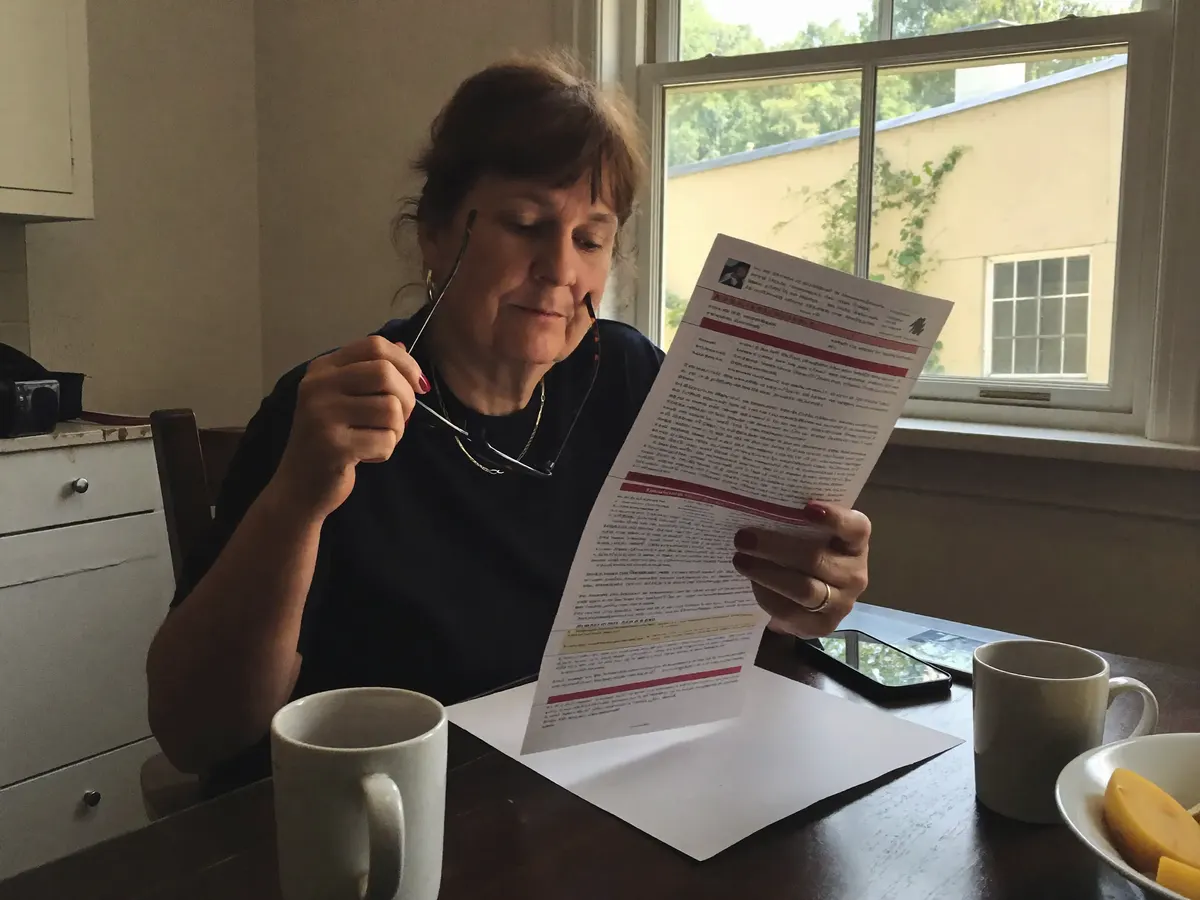 A woman reads a printed agreement at her kitchen table through reading glasses.